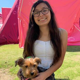 Citlali is sitting on the grass in a white sweetheart neckline buttoned tank top and high waisted blue jeans. Bubbles are in the air. She is wearing her glasses and her long, brown hair is down. She is in front of a large pink tent and is holding her dog Chewie, a brown Yorkshire Terrier with a green bow.