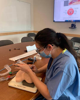 Citlali has her long brown hair tied back in a pony tail and is wearing a surgical mask. She is pictured in side-profile sitting in blue scrubs. She is practicing intubation on a mannikin head in a conference room at Brigham and Women's Hospital Neonatal Intensive Care Unit.