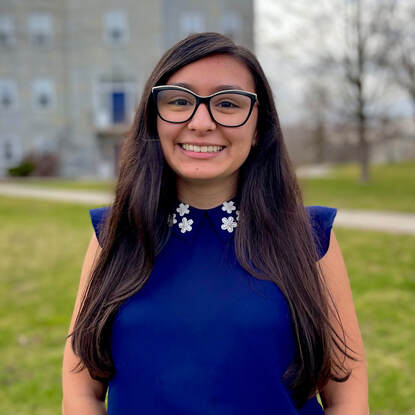 Citlali has brown eyes, long brown hair, and is tan skinned. In this head shot she is smiling wide and is wearing glasses and a dark blue blouse with a white flowered collar. She is standing in front of the bright green lawn in front of Old Chapel.